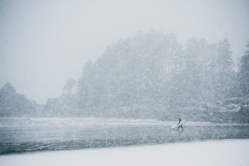 A surfer walking through the snow on their way out to the water to surf in Tofino in winter conditions